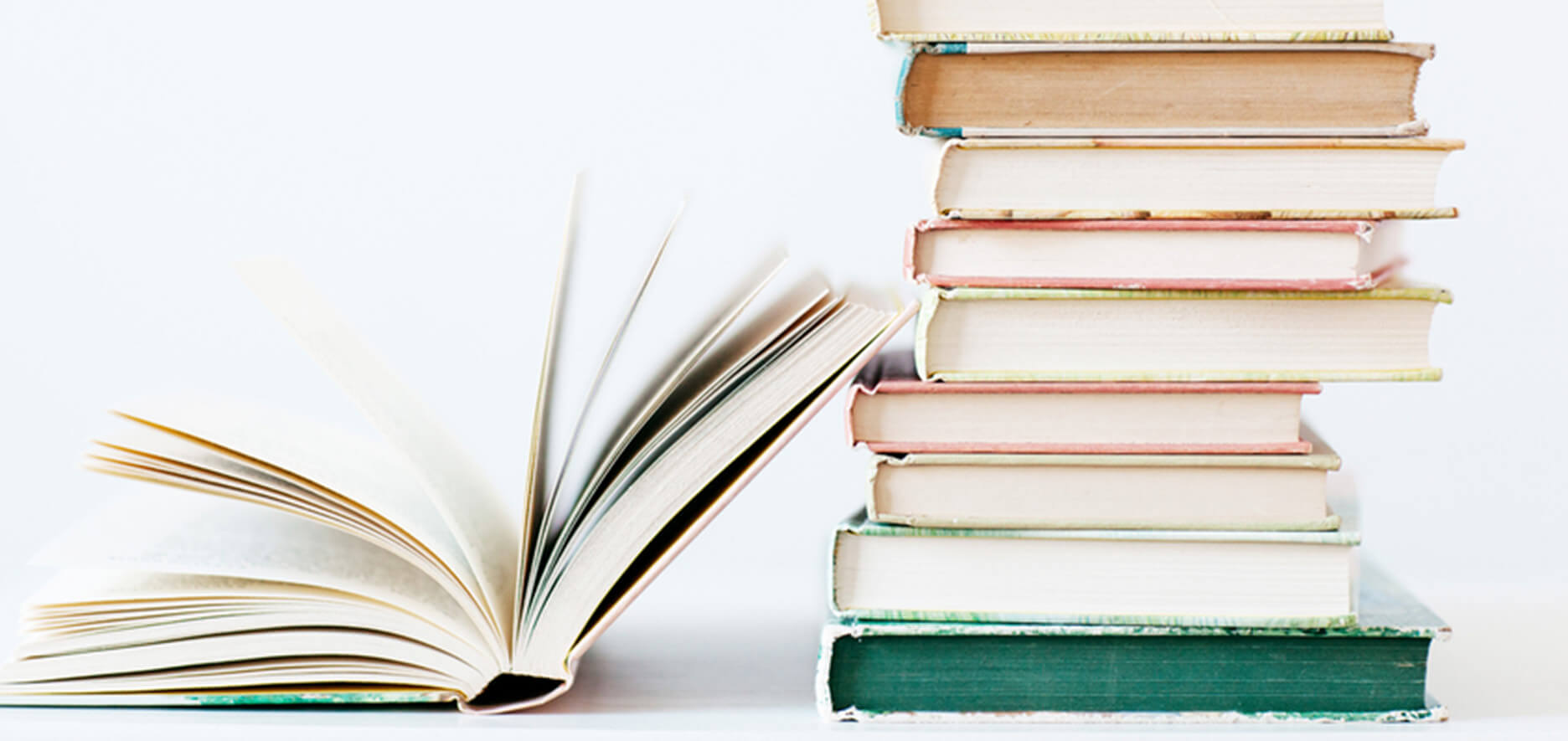 A Stack Of Nine Books Stands Next To An Open Book On A White Surface Against A Plain White Background.