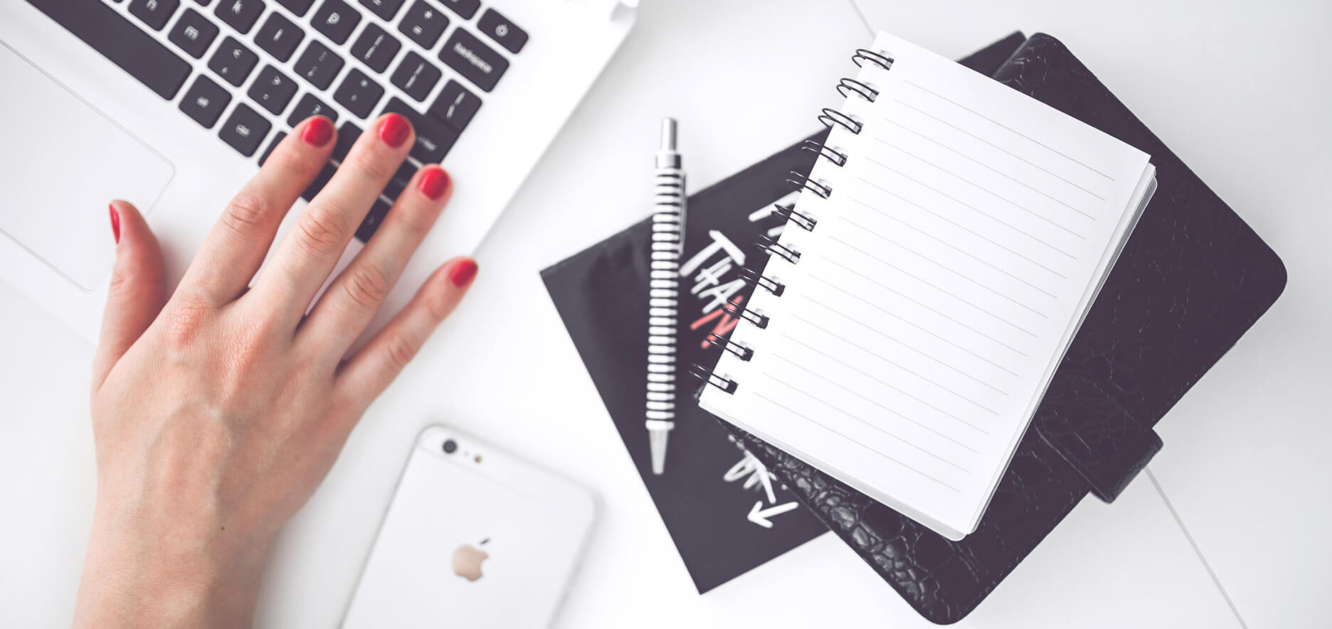 Hand With Red Nail Polish Typing On A Laptop Next To A White Smartphone, A Spiral Notebook, And A Black Leather-Bound Planner.