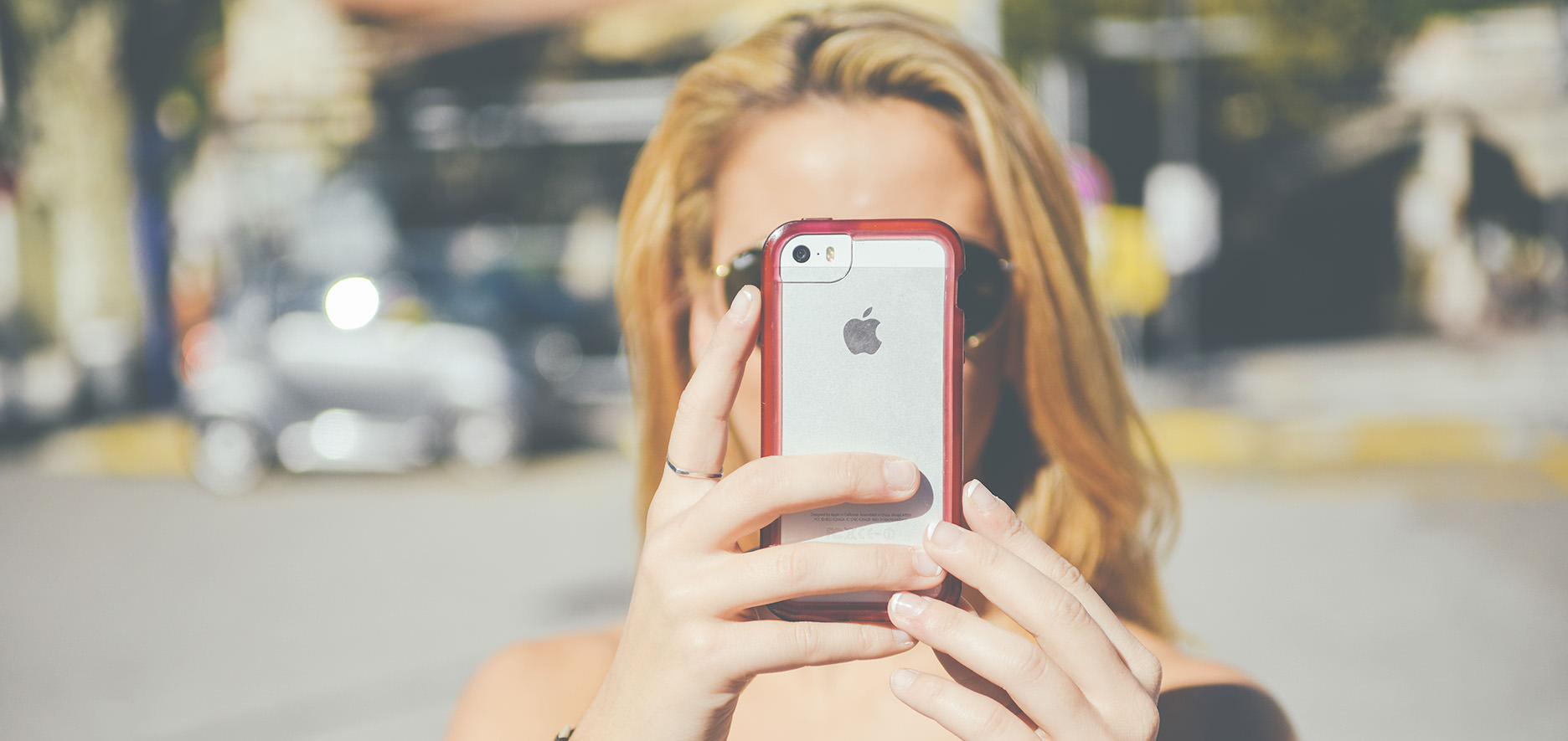 A Person With Blonde Hair Holds Up A Silver Iphone With A Red Case, Facing The Phone'S Screen. The Background Is Blurry, Showing Outdoor Scenery.