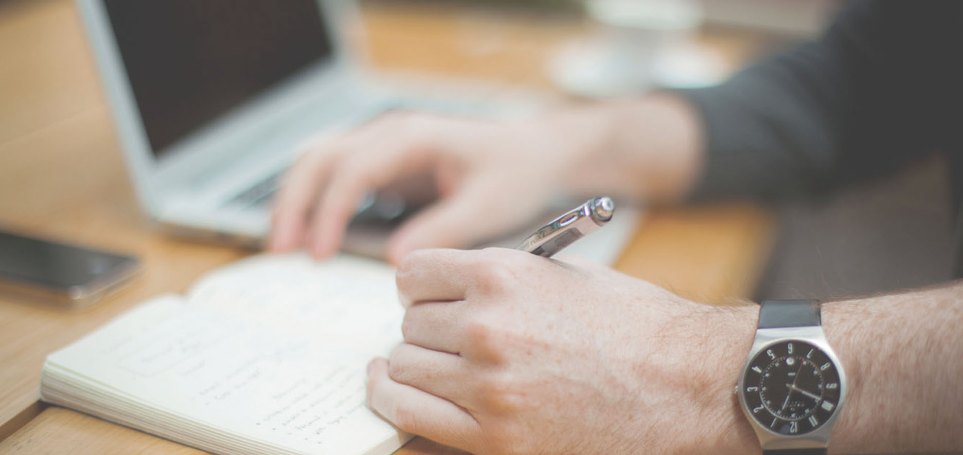 A Person Wearing A Wristwatch Writes In A Notebook With A Pen Next To An Open Laptop On A Wooden Desk.