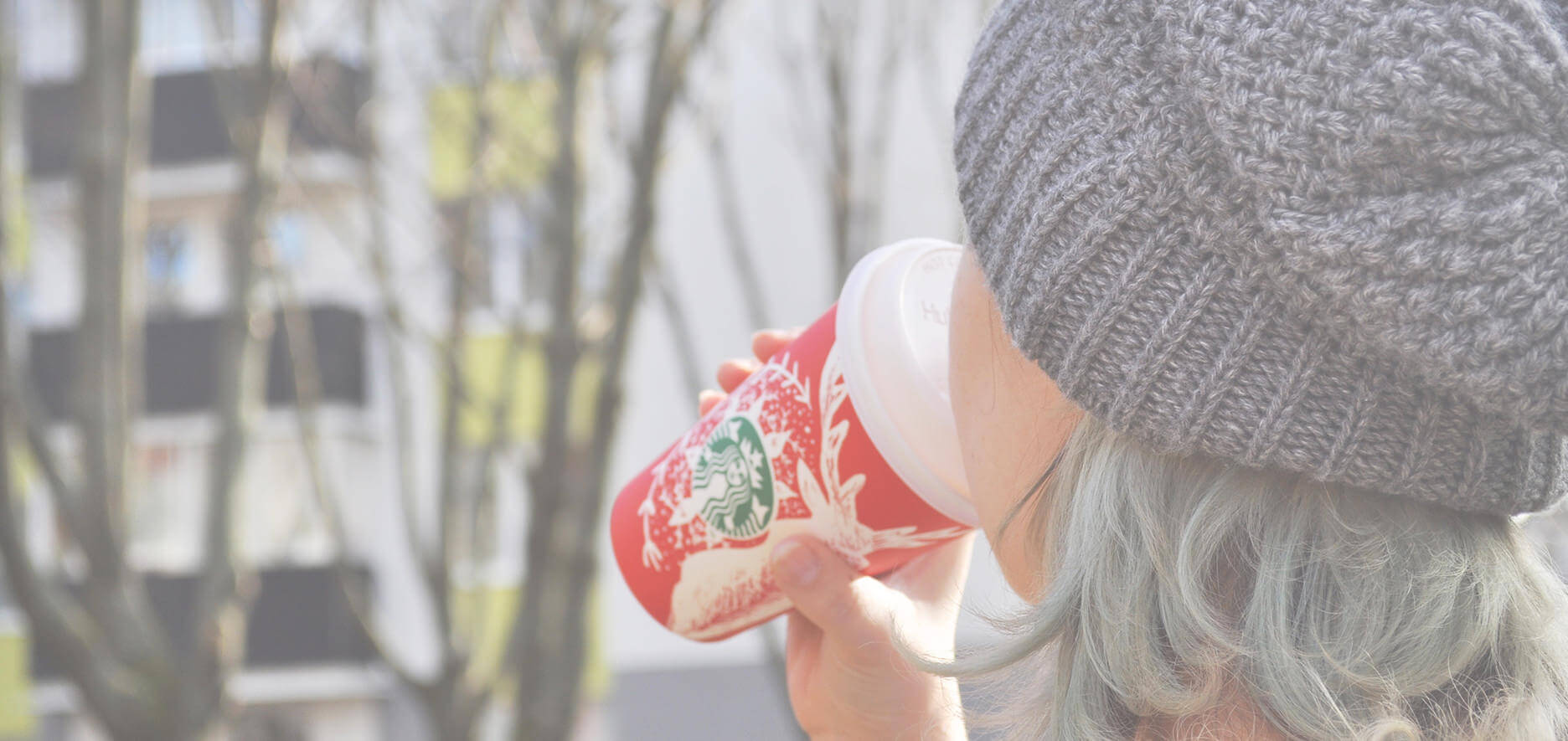 A Person Wearing A Gray Knit Hat Drinks From A Red Starbucks Cup Outdoors With Trees And Buildings In The Background.