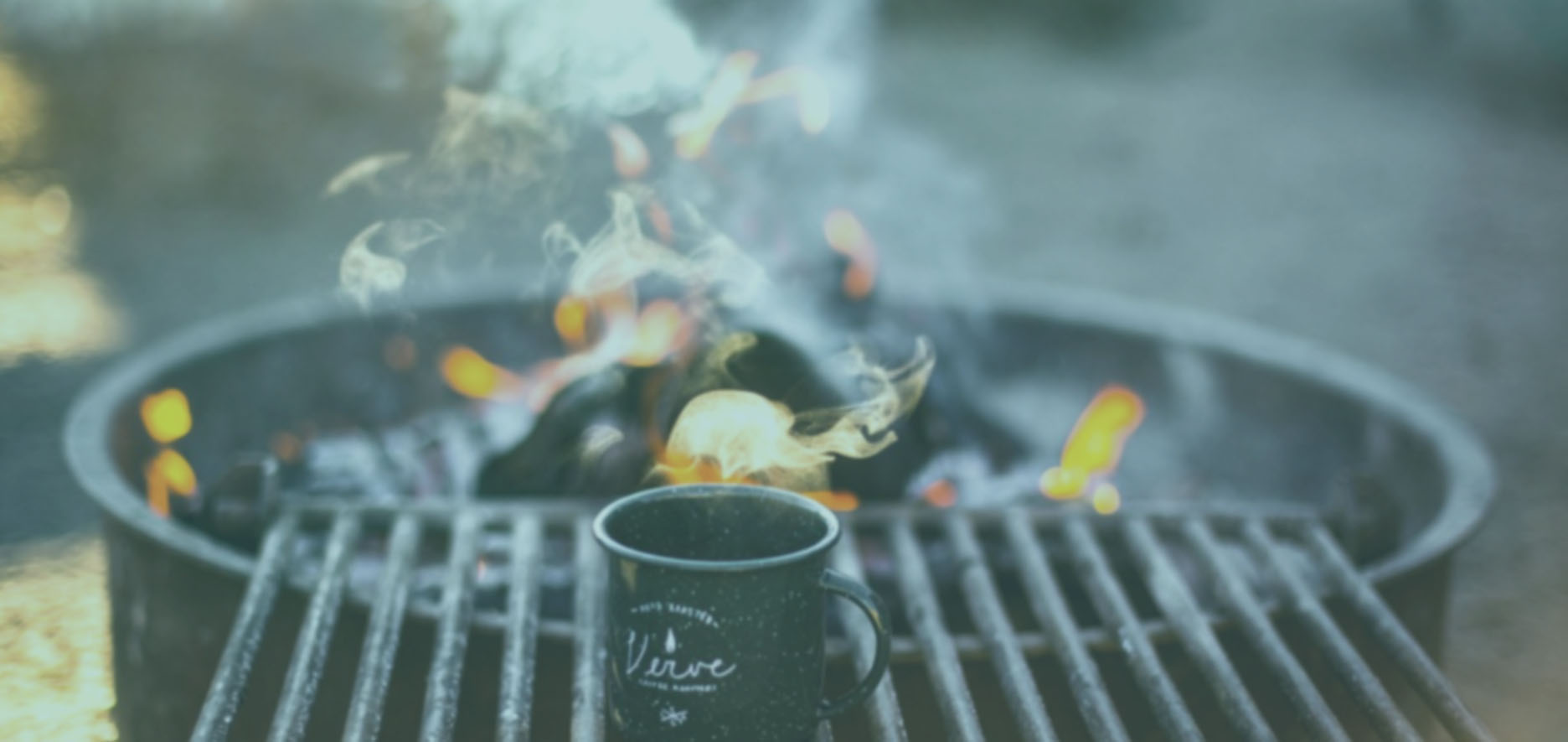 A Steaming Mug Sits On A Metal Grill Near A Campfire With Visible Flames And Smoke.