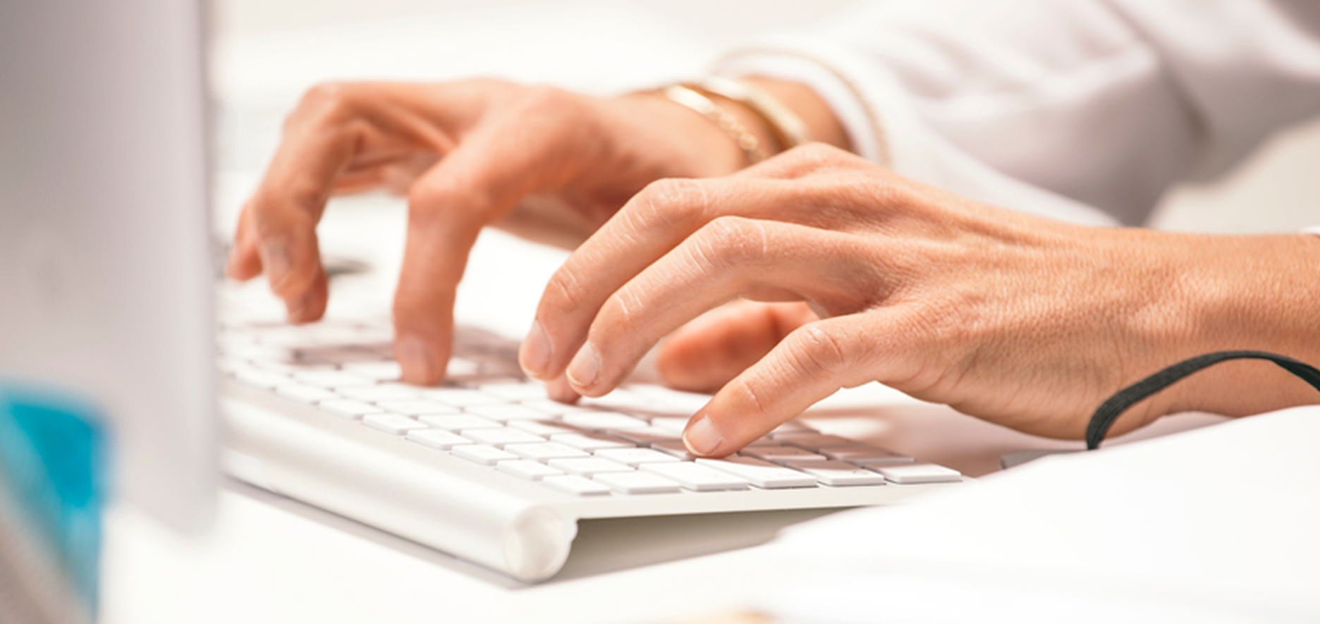 Hands Typing On A White Keyboard With A Wristwatch Visible On The Left Wrist. The Background Is Blurred.