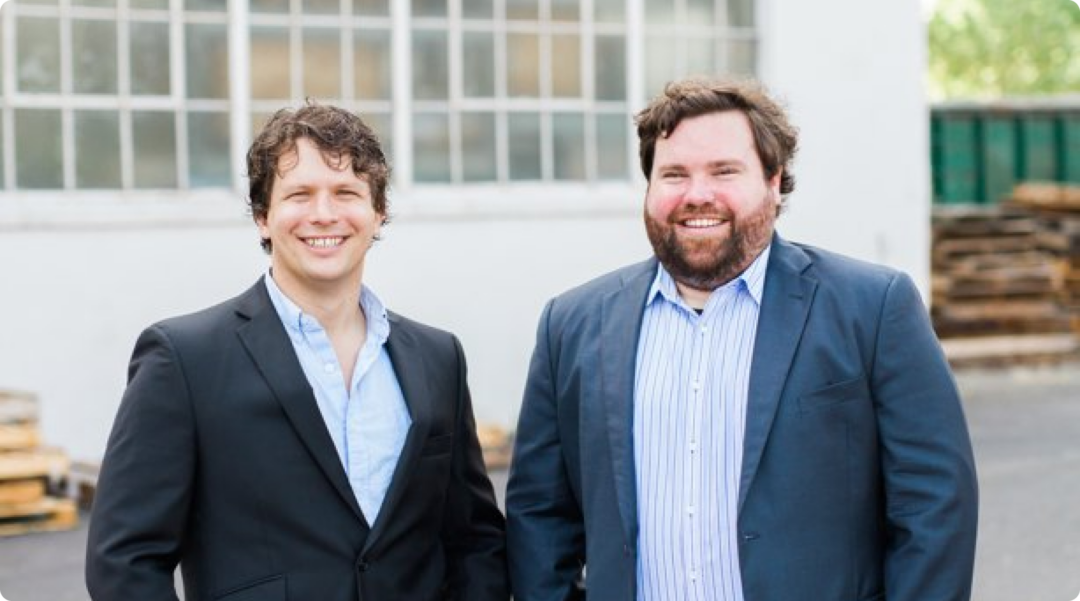 Two Men, Both Wearing Suits And Smiling, Stand Outdoors In Front Of A Building With Large Windows.