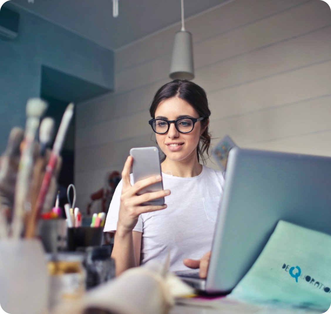 A Person Wearing Glasses Sits At A Desk With A Laptop And Art Supplies, Looking At A Smartphone.